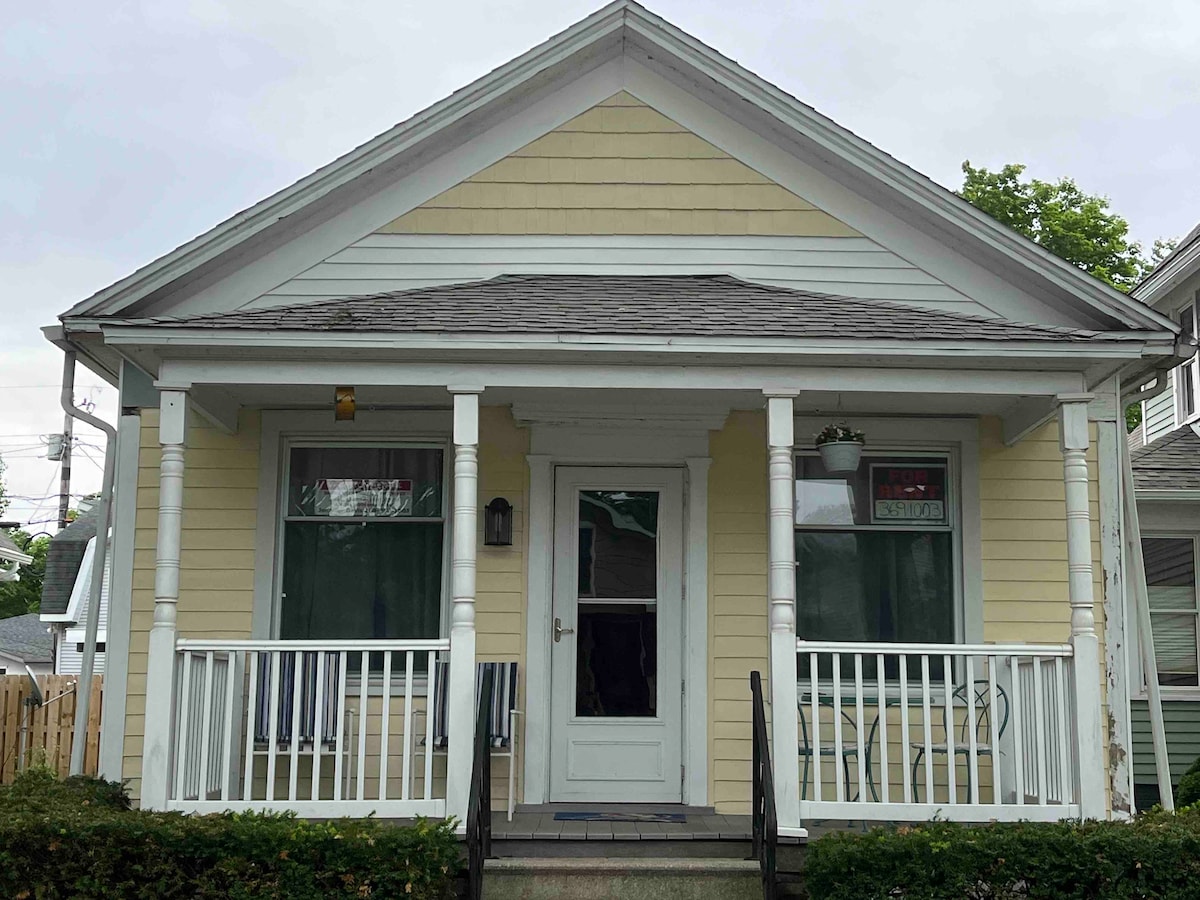 The front of a two-bedroom house features a welcoming porch with white railings and two entry steps. Symmetrically placed windows are framed with green curtains, and a door is centrally located. The exterior is painted a soft yellow, contributing to the home's charm.
