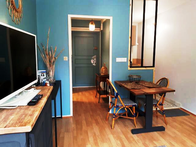 A bright living space is presented with a blue accent wall. A flat-screen TV is mounted on a wooden console, while a dining table with chairs is visible. The entrance features a green door, enhancing the inviting ambiance of the area.
