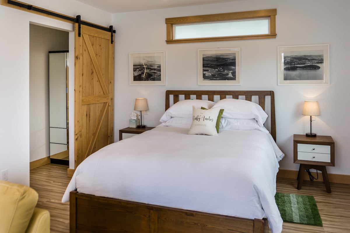 A spacious sleeping area features a wooden bed adorned with white bedding and decorative pillows. Two bedside tables with lamps flank the bed, while framed photographs hang on the walls. A sliding barn door leads to a mirrored closet, enhancing the modern aesthetic.