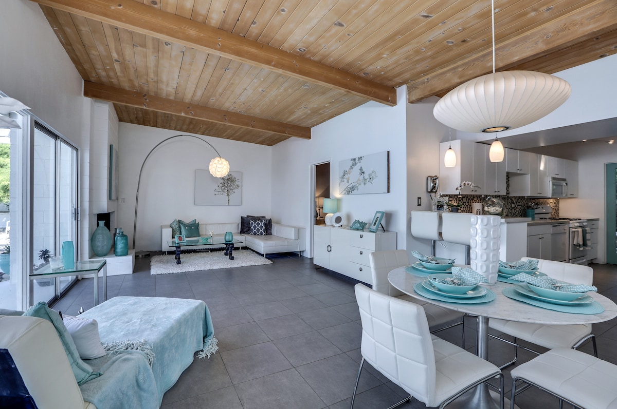 A spacious living area features a light-colored wood ceiling and modern furnishings in soft blue tones. A round dining table is set with turquoise dishes, while a comfortable seating area is arranged with white sofas and decorative accents. Natural light streams through large glass doors.
