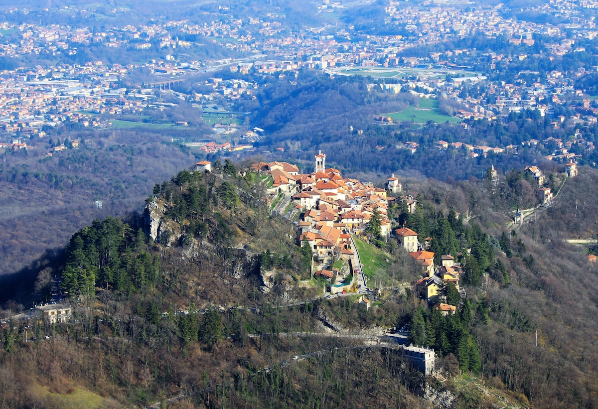 A panoramic view of a hilltop village, showcasing terracotta-roofed buildings nestled among trees. The landscape transitions into valleys and distant mountains, with visible urban areas spreading across the horizon under a clear blue sky.