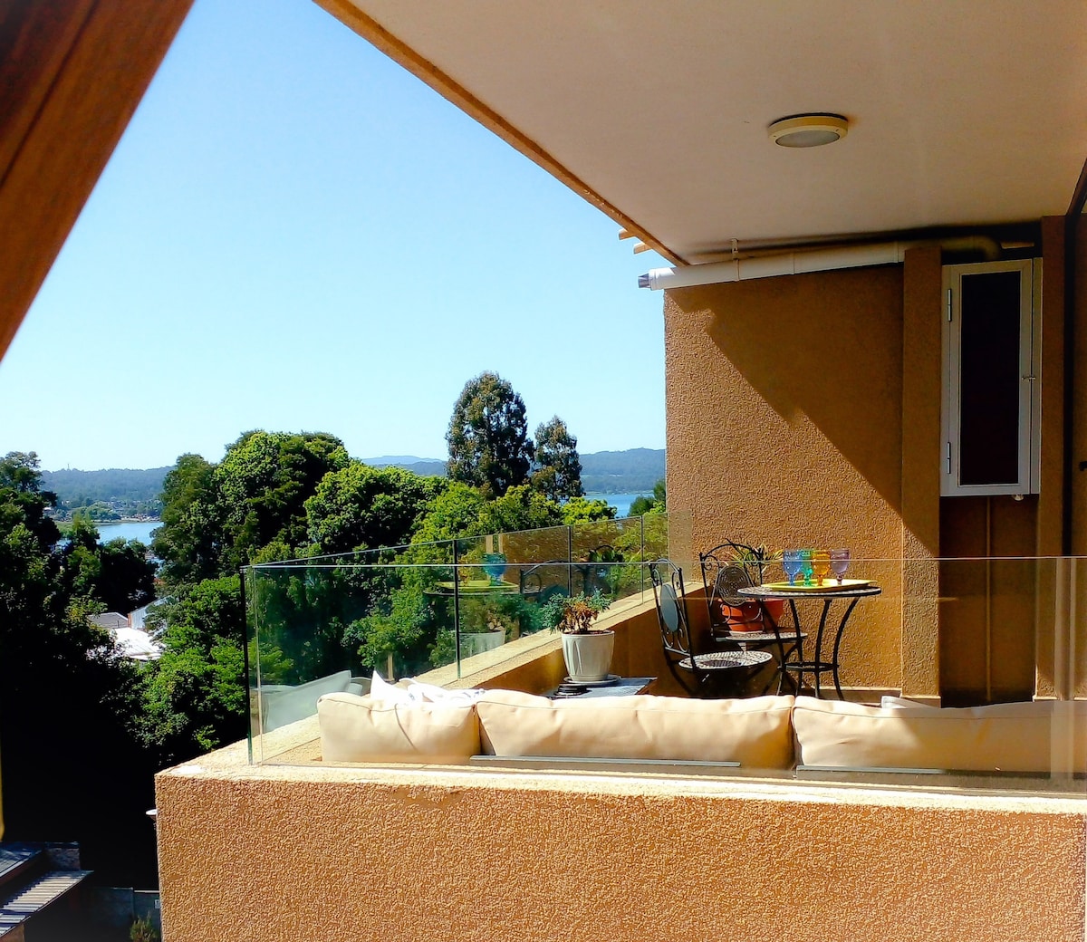 A terrace features comfortable seating with soft cushions and a small round table. Lush greenery surrounds the area, providing a serene view of the lake in the distance under a clear blue sky.