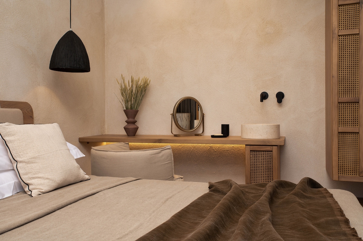 A serene bedroom is presented, featuring a spacious bed with neutral linens. A minimalist table with a round mirror and decorative elements is positioned against a textured wall. Soft lighting highlights the details, creating a relaxing ambiance in this inviting space.