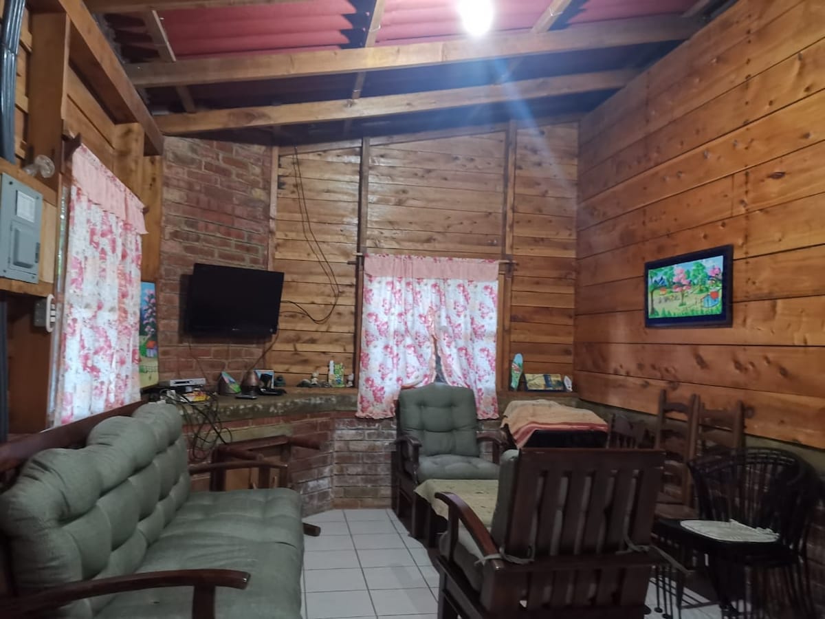 A cozy living area features wooden walls and ceilings, complemented by large windows adorned with floral curtains. Comfortable seating arrangements include a sectional sofa and armchairs. A mounted television is visible, and artwork hangs on the wall, enhancing the ambiance.