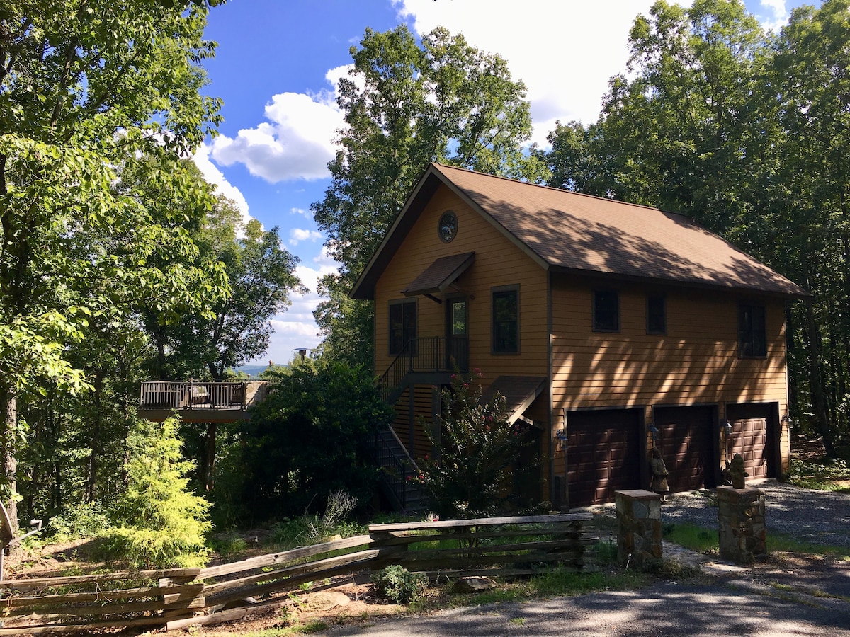 A two-story wooden house is surrounded by greenery, with a driveway leading up to the entrance. A deck is visible on the left side, offering a vantage point of the landscape. The entrance features a staircase leading up to the main level.