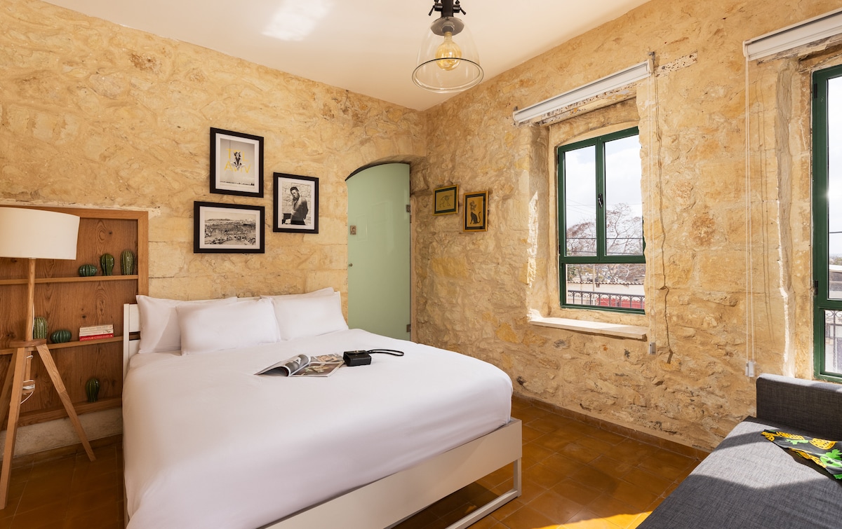 A bright bedroom features natural stone walls and large windows that allow light to enter. A spacious bed is positioned against the wall, complemented by a bedside table and a stylish lamp. Framed artwork adds character to the inviting space.