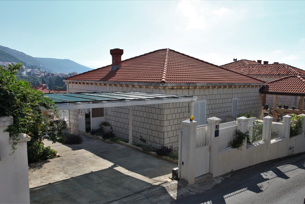 A spacious family house is depicted, featuring a traditional red-tiled roof and light-colored stone walls. A covered area provides shaded parking access. Surrounding greenery is visible, contributing to a peaceful atmosphere. The nearby hills create a serene backdrop against the clear sky.
