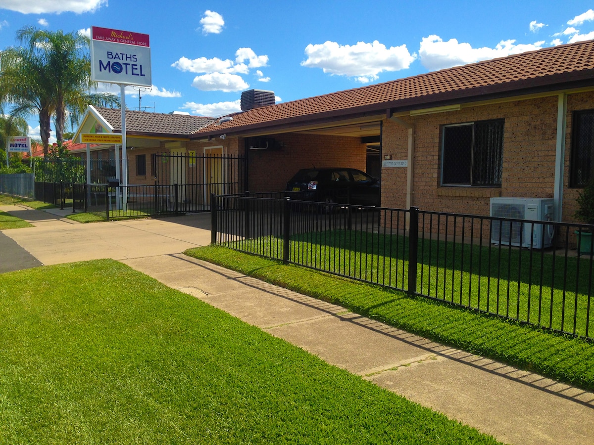 The exterior of Baths Motel is shown, featuring a long, brick building under a clear blue sky. A well-maintained lawn with green grass encircles the property. Parking spaces are available next to the entrance while the motel sign is clearly visible.