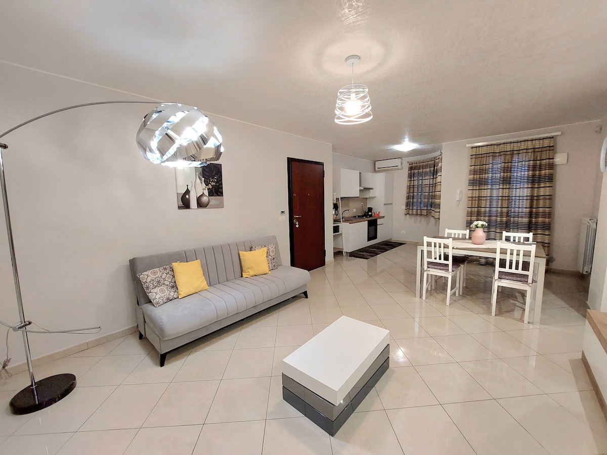 The living area features a light, modern design with a gray sofa adorned with two yellow cushions. A white coffee table is positioned in front of the sofa. A dining table with white chairs is visible, accompanied by a well-equipped kitchen in the background.