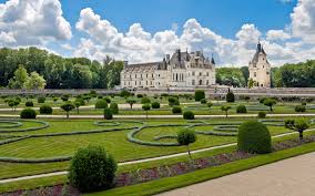 A historical château is seen in the background, surrounded by neatly manicured gardens. The landscape features symmetrical hedges shaped into patterns and a variety of greenery under a bright blue sky dotted with fluffy clouds.