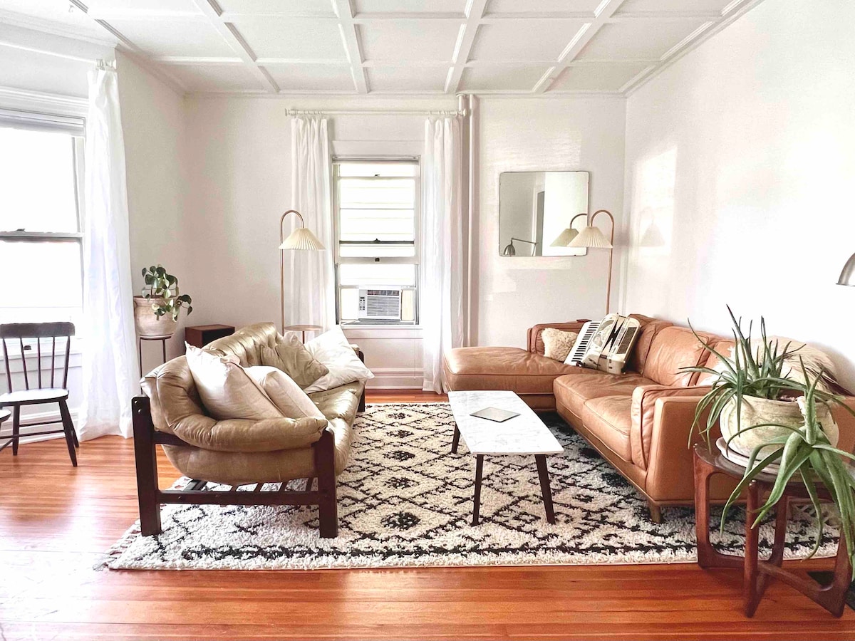 A cozy living area features a leather sectional sofa alongside a plush, oversized armchair. A low coffee table is positioned on a patterned area rug. Light streams through large windows adorned with sheer curtains, creating a warm and inviting atmosphere in the space.
