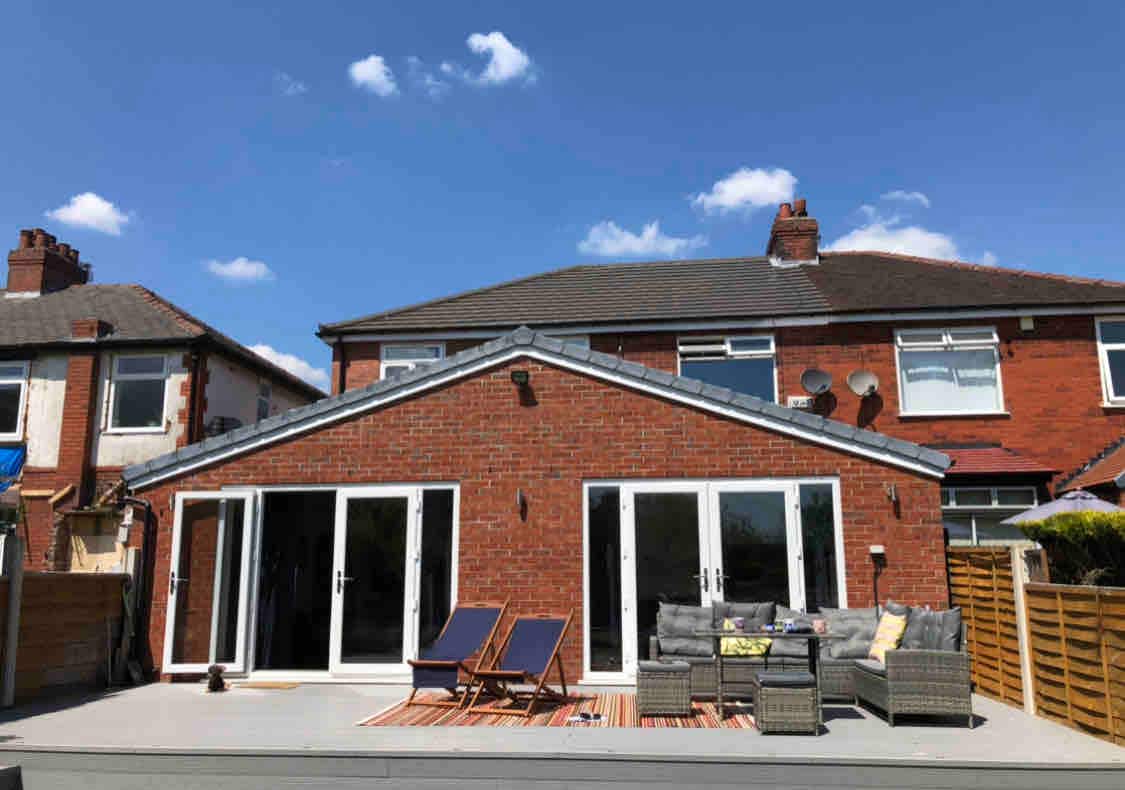 The rear of the property features a spacious patio area with modern outdoor furniture, including lounge chairs and a seating arrangement. Large sliding glass doors provide access to the interior, while the sky above is clear and blue, enhancing the inviting outdoor space.
