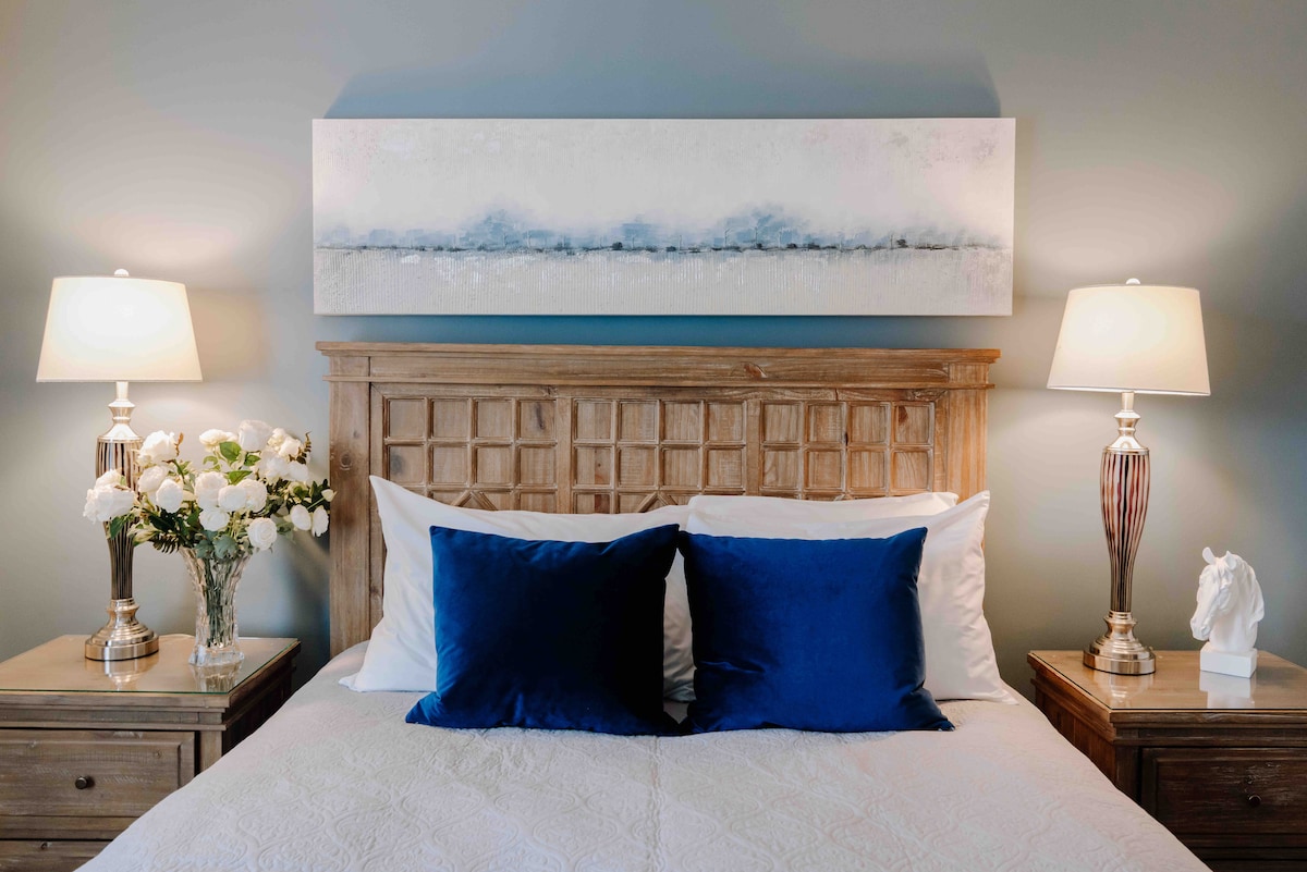 A well-appointed bedroom features a wooden headboard above a spacious bed with white linens and deep blue accent pillows. Nightstands with lamps flank each side, and a piece of abstract art in soft hues hangs above the bed. A bouquet of white flowers adds a touch of elegance.