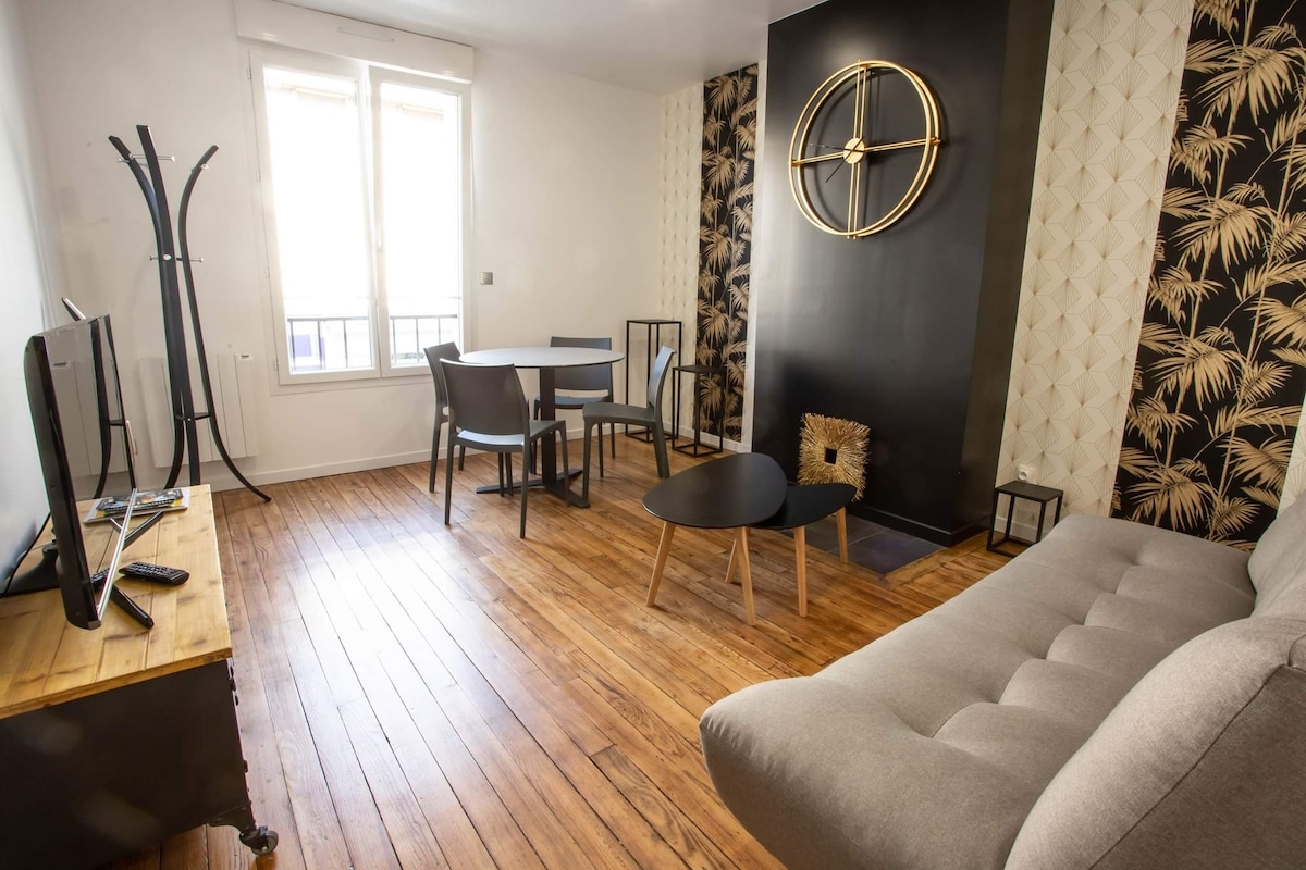 A stylish living area features hardwood flooring and decorative wall accents. A comfortable sofa faces a modern television. A small dining table with four chairs occupies one corner, alongside large windows allowing natural light to fill the room. A decorative wall clock adds character.