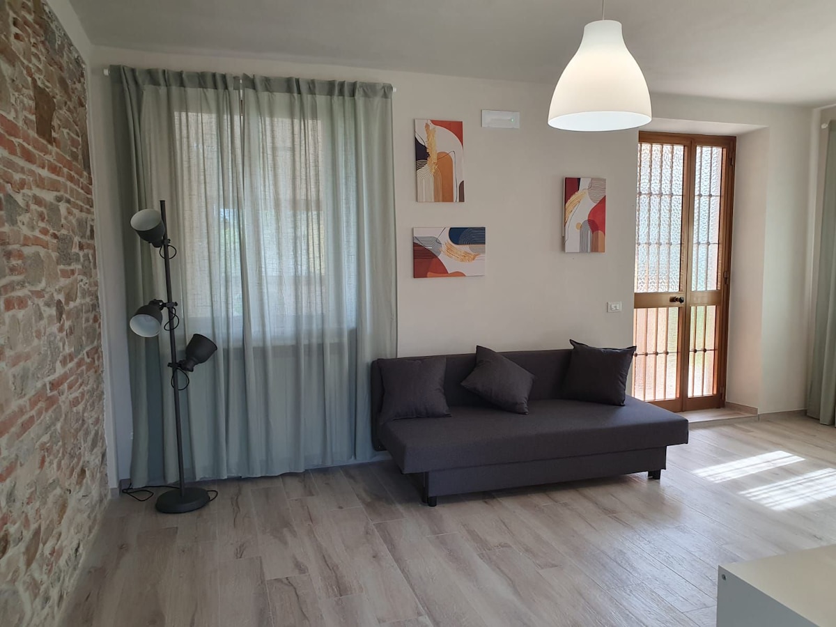 A bright living area features a modern gray sofa adorned with decorative pillows. Natural light enters through a large window, softly illuminating the space. Complementary wall art adds visual interest against a backdrop of exposed brick and neutral wall colors.