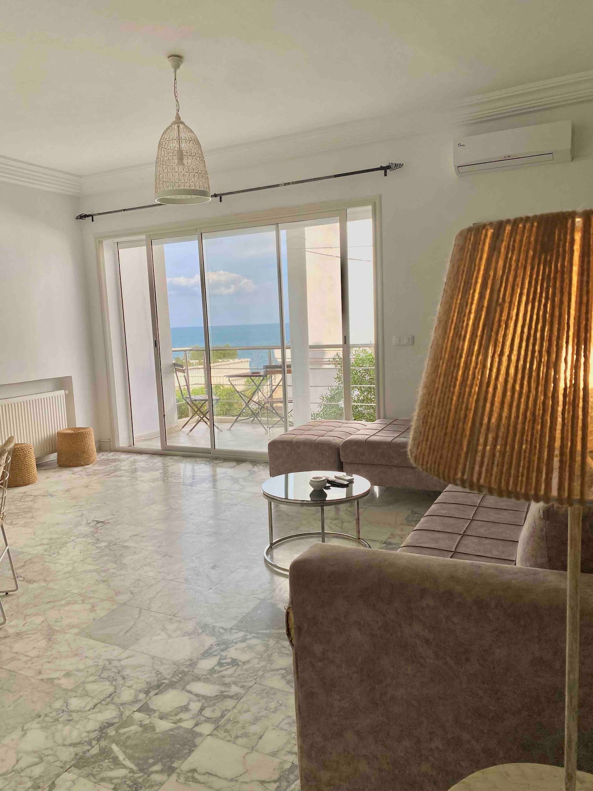 A spacious living area is highlighted by large glass doors offering a view of the sea. The room features light marble flooring, a low coffee table, and a cozy seating arrangement. A stylish lamp adds a touch of warmth to the space.