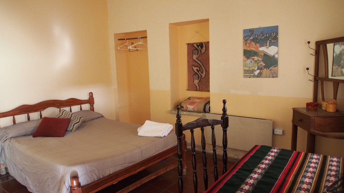 A spacious room features a wooden bed with neutral bedding and decorative pillows, complemented by a small table and chair. A wall-mounted window showcases a scenic view. Decorative textiles add visual interest, while a wardrobe and hooks provide convenient storage options.