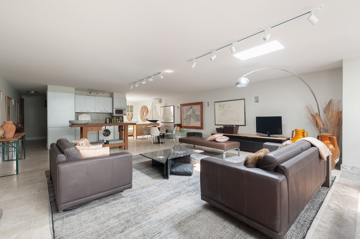 An open-plan living area features two dark leather sofas facing a glass coffee table. A sleek kitchenette is visible in the background, complemented by dining seating. Light is provided by overhead fixtures and a skylight, creating an airy atmosphere. Decorative accents and artwork adorn the walls.