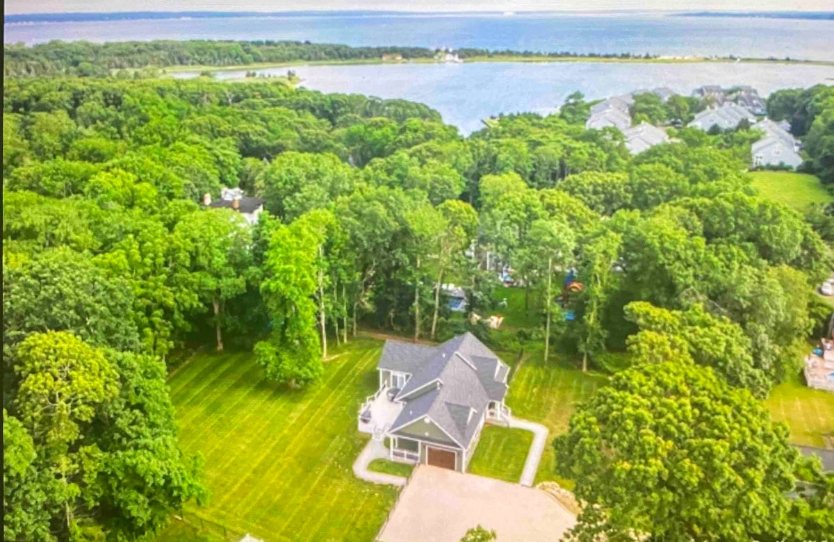 An aerial view captures a newly constructed beach house nestled within lush greenery. The property is surrounded by manicured lawns, with a glimpse of a serene body of water in the background. Tree canopies provide a natural frame to the inviting space.