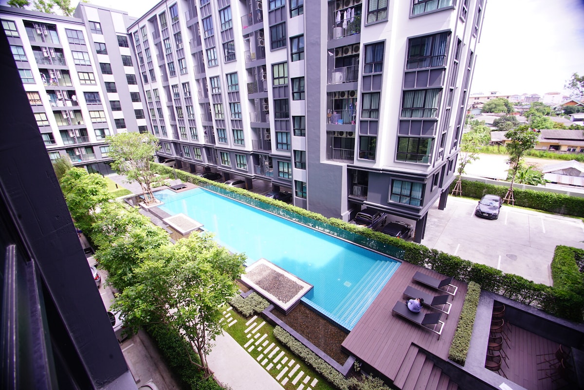 An aerial view of the outdoor swimming pool is presented, surrounded by greenery and lounge chairs. The modern condo building features large glass windows, reflecting natural light. The pool area is designed with minimal landscaping, ensuring an inviting space for relaxation.