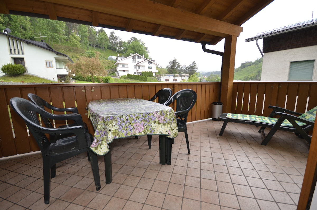 A spacious veranda features a table covered with a floral cloth, surrounded by four black chairs. A lounge chair is positioned nearby, complemented by tiled flooring and wooden beams overhead, providing an inviting area for outdoor dining and relaxation.