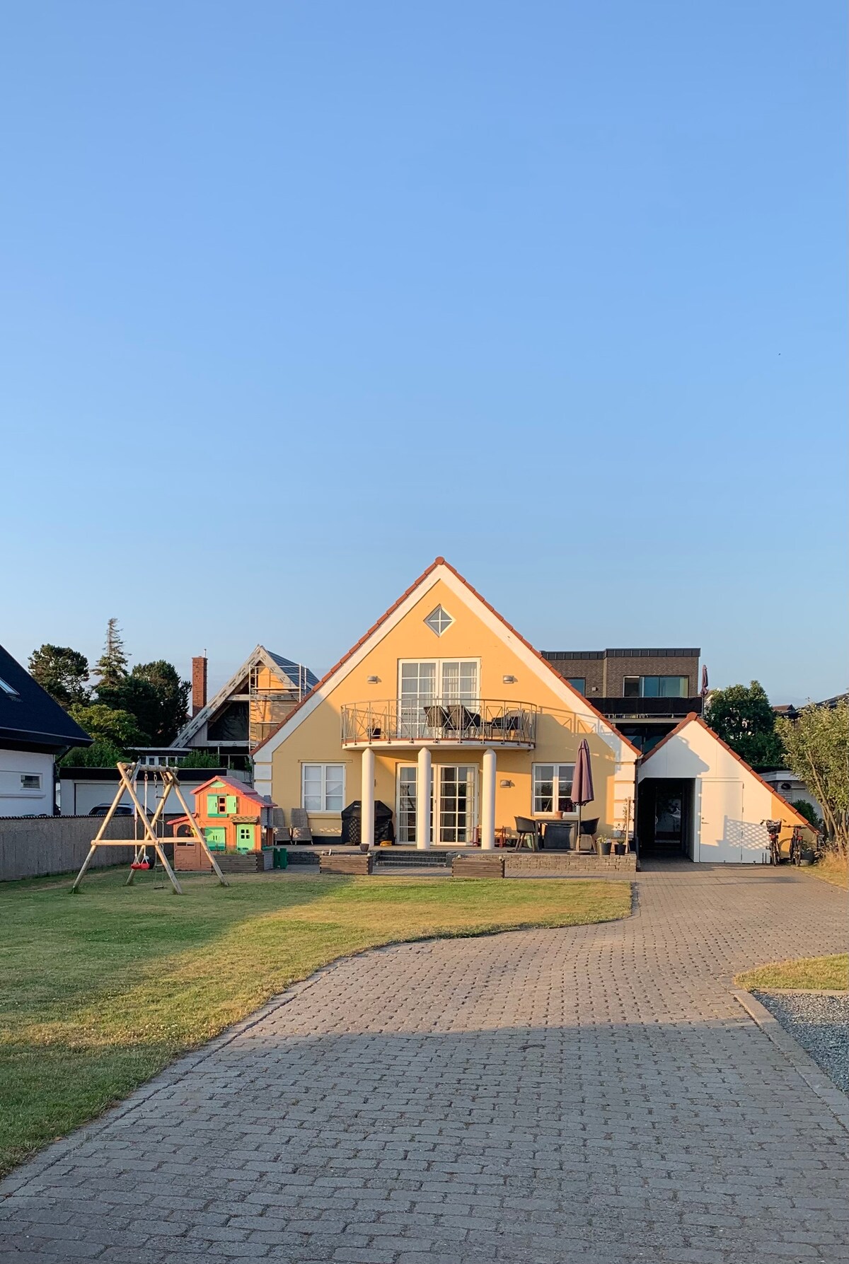 A charming, yellow house features a pitched roof and multiple balconies. Lush green lawn surrounds the structure, complemented by a stone pathway leading to the entrance. A playground structure with swings is visible in the yard, adding to the welcoming outdoor space.
