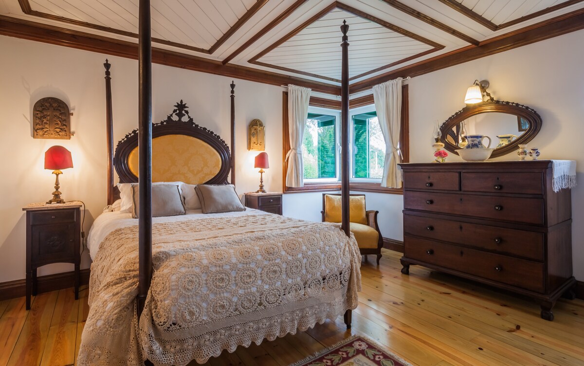 A well-appointed bedroom features a four-poster bed with an ornate headboard and soft bedding. Two bedside tables with red lamps frame the bed, and a wooden dresser stands against the wall. Natural light enters through a green-trimmed window, complemented by elegant curtains.