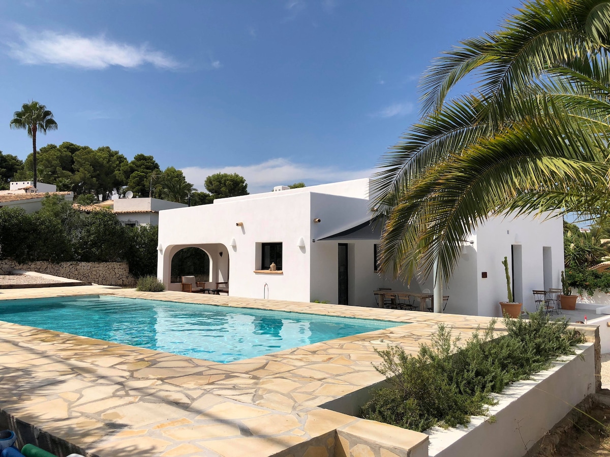 The exterior of a modern villa is shown, featuring a private outdoor pool surrounded by a stone patio. Lush greenery, including palm trees, is visible in the background, contributing to a serene environment. Large windows reflect natural light, enhancing the villa's spacious feel.