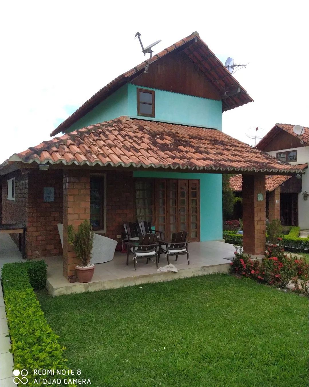 A two-story house features a rustic brick exterior with a blue accent wall. A covered porch with a set of four chairs overlooks a well-kept lawn, bordered by manicured bushes and colorful flower beds.