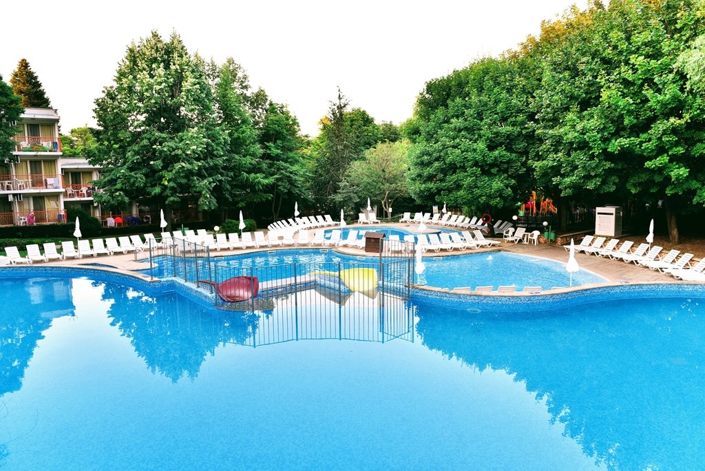 A large outdoor swimming pool is surrounded by lush greenery, providing a serene environment. Lounge chairs are arranged neatly along the pool's edge. A safety fence encloses a shallow play area within the pool, ensuring a safe space for children.