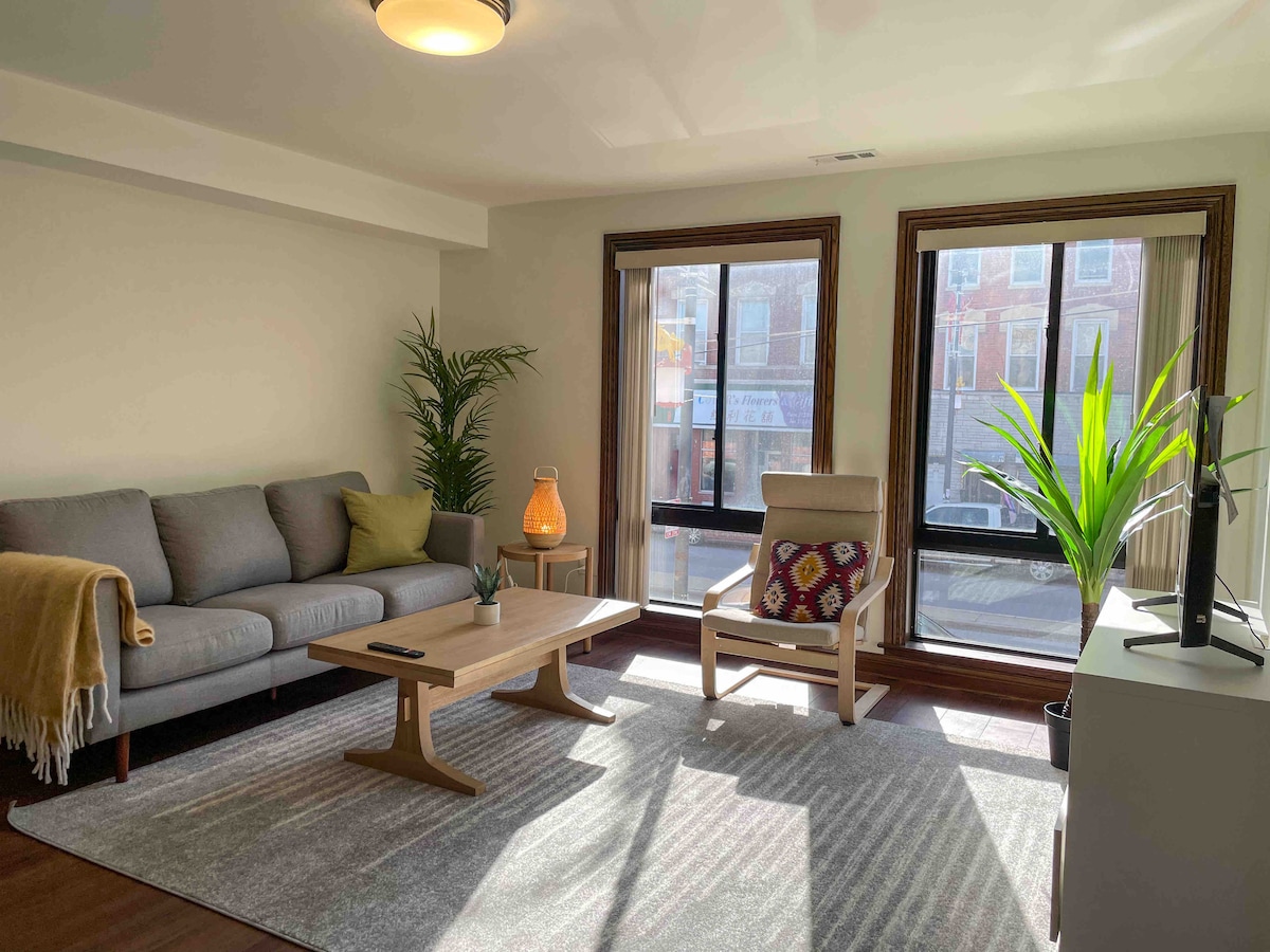 A bright living area is showcased with a grey sofa and a wooden coffee table. Large windows allow natural light to fill the space, complemented by indoor plants. A textured rug lies beneath, and a small decorative lamp adds a touch of warmth.