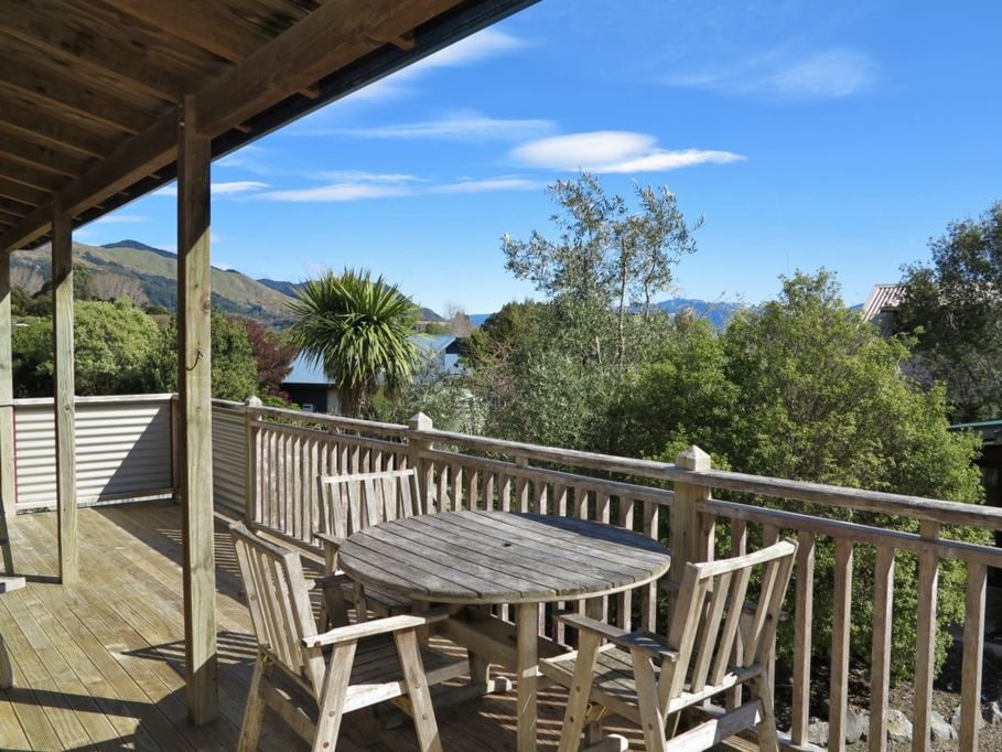 A spacious outdoor deck features a round wooden table surrounded by four matching chairs, offering a tranquil view of the surrounding greenery and distant mountains. Bright blue skies complement the serene natural setting.
