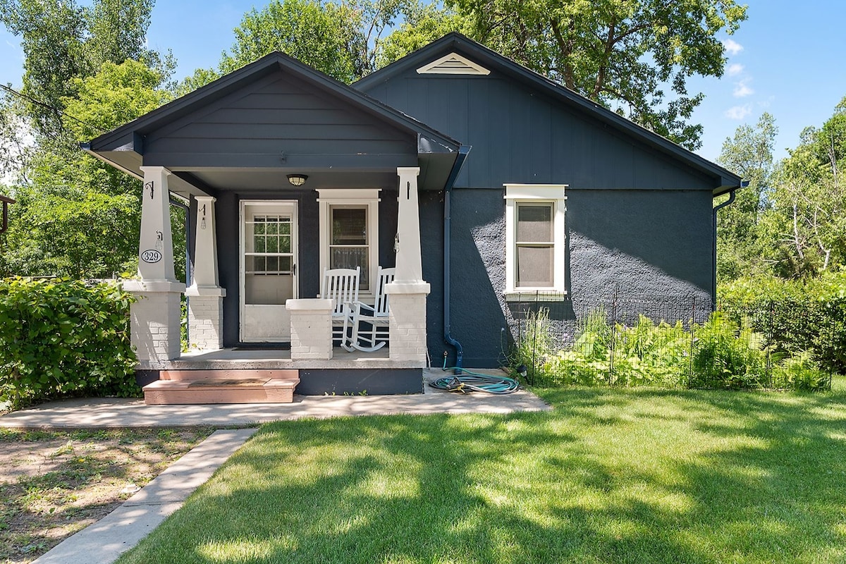 The exterior of a welcoming home is shown, featuring a covered porch with two white rocking chairs. Lush green grass surrounds the pathway leading to the entrance, while vibrant greenery lines the sides of the house. Natural light illuminates the front, enhancing its charm.