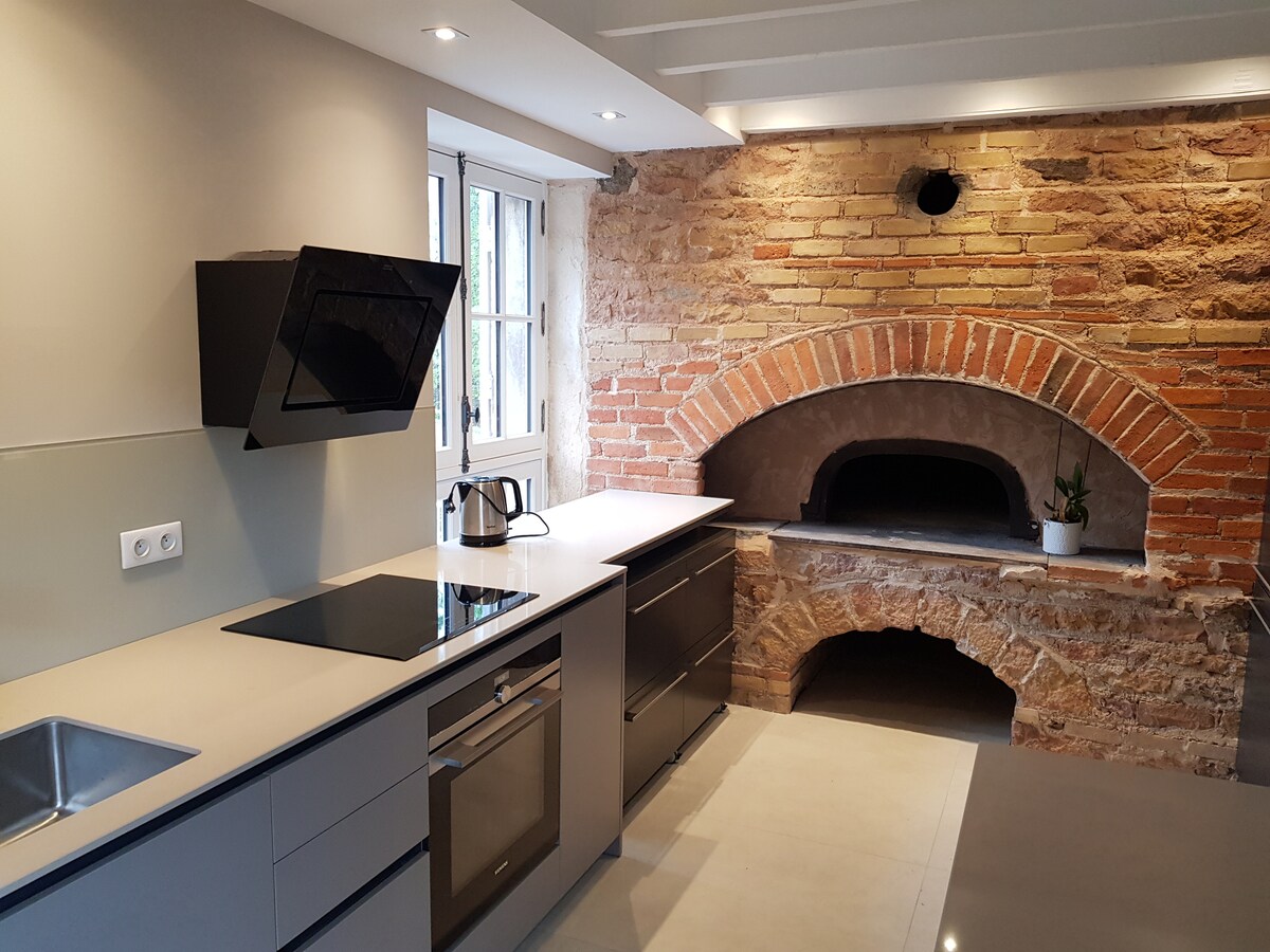 The modern kitchen features sleek cabinetry and an integrated stove. A rustic brick oven is positioned prominently against the wall, showcasing its textured surface. Natural light filters through the window, illuminating the spacious countertop and minimalist design.