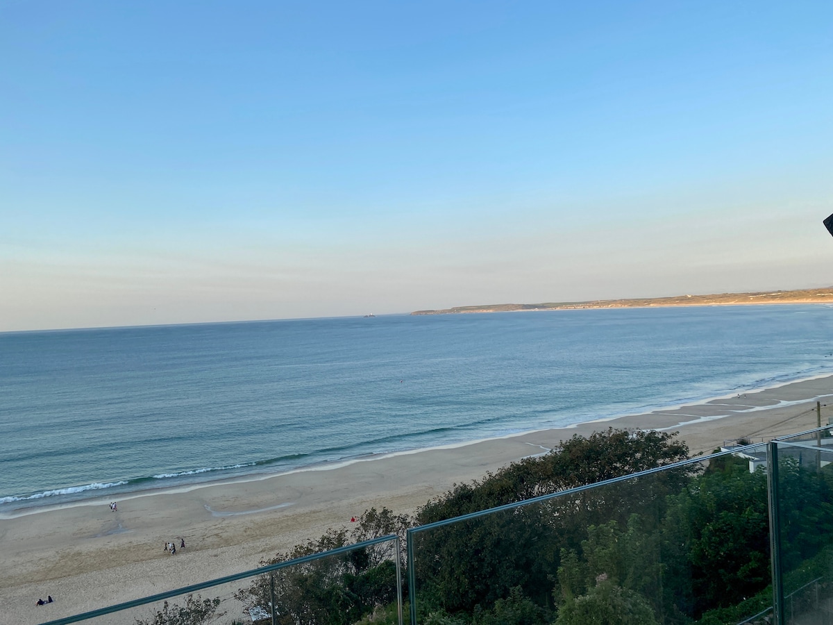 A panoramic view of Carbis Bay Beach is visible, featuring soft sandy shores and gentle waves. The horizon blends into the calm sea under a clear blue sky, with distant coastal outlines fading into the background.