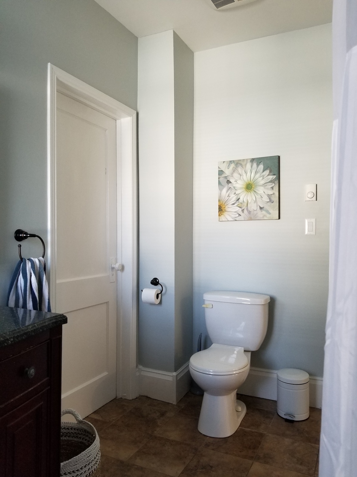 A well-lit bathroom features a toilet adjacent to a decorative flower wall art piece. A dark wooden vanity with a granite countertop is present, along with a waste bin and towel holder. Light blue walls create a calming atmosphere.