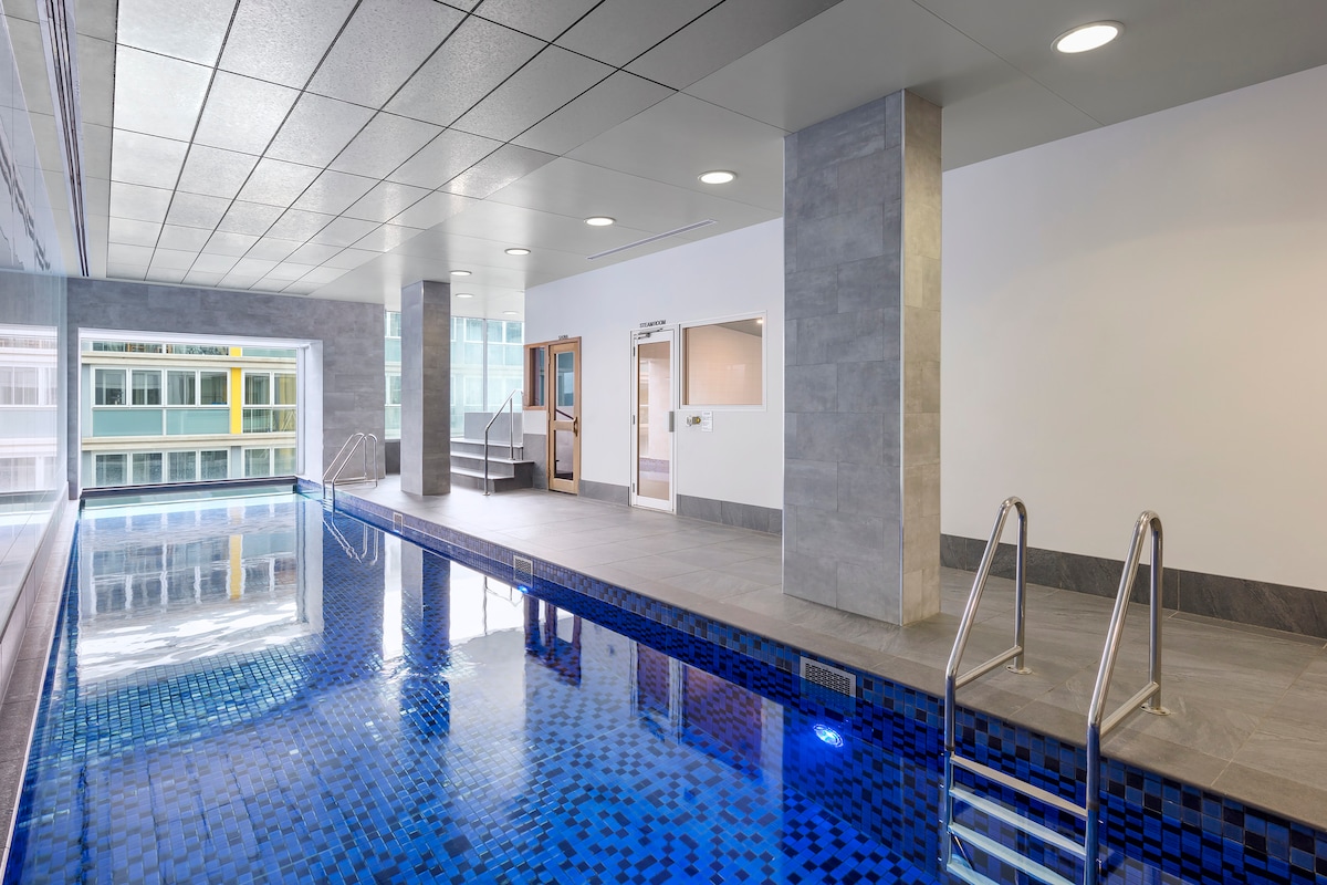An indoor heated pool is presented with clear blue water reflecting overhead lights. The area features modern, smooth gray tiles and large windows, allowing for natural light. Ladder steps lead into the pool, and adjacent spaces are visible, providing access to changing facilities.