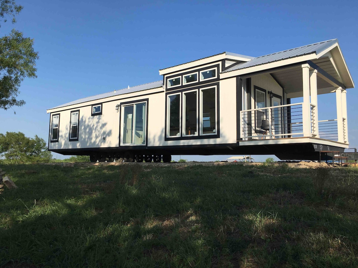 A modern house is depicted, showcasing large glass windows that allow natural light to fill the interior. A spacious porch extends out, offering an outdoor seating area. The structure is elevated on supports, surrounded by green grass and trees, set against a clear blue sky.