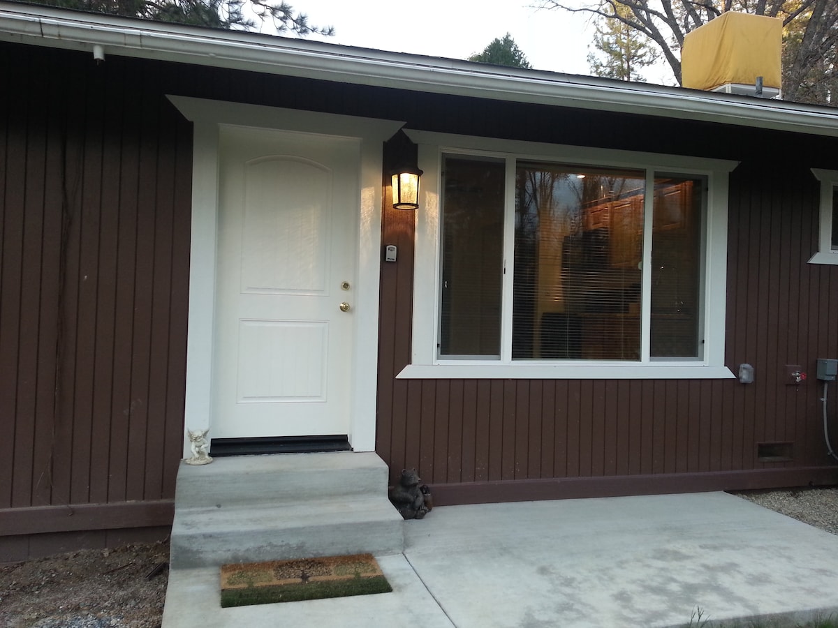 The entrance features a white front door with a modern handle, flanked by large windows allowing natural light. A concrete step leads up to the door, while a small doormat is positioned in front. A light fixture is mounted beside the door.