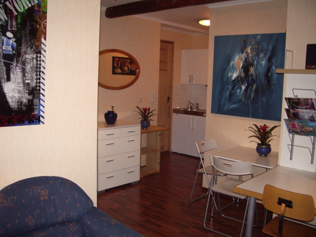 An inviting living space is depicted, featuring wooden flooring and light-colored walls. A blue sofa is positioned near a contemporary wall art piece. A dining table with chairs sits adjacent to a kitchenette, which includes a refrigerator and cabinets, while decorative plants add a touch of greenery.