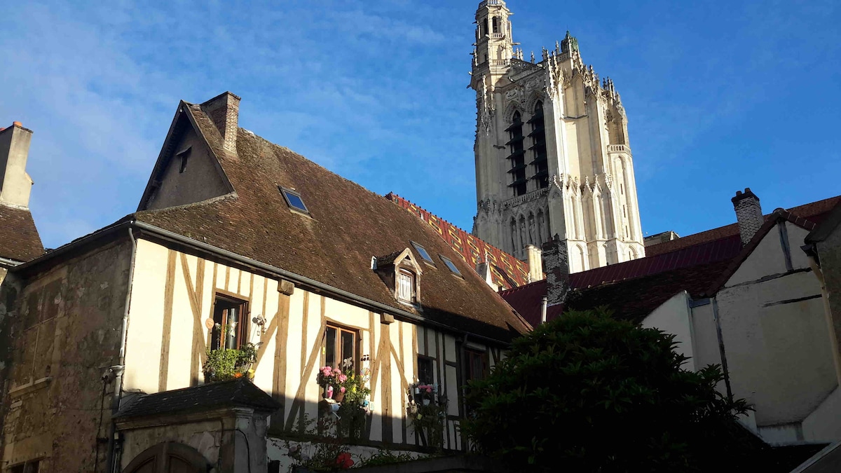 The image captures a historic building with traditional timber framing in the foreground, flanked by vibrant flowers. A towering church spire rises majestically in the background, set against a clear blue sky, highlighting the charming architecture of the area.