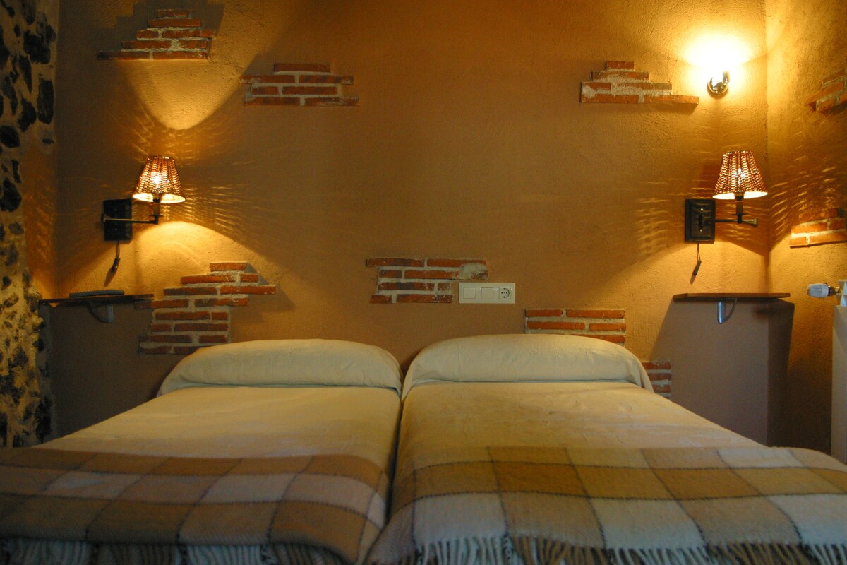 A warm and inviting sleeping area features two separate beds with light-colored bedding. The walls are adorned with exposed brick accents and gently illuminated by bedside lamps. Shelves are situated on either side of the beds, enhancing the room's cozy design.