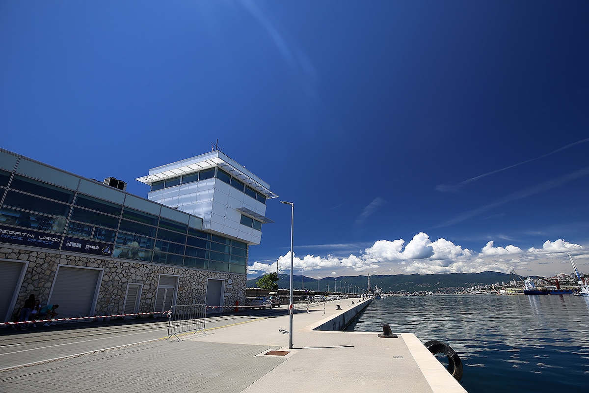 A modern waterfront building features large glass panels that reflect the vibrant blue sky. The well-maintained promenade extends along the water's edge, offering a scenic view of the harbor and distant clouds. The expansive shoreline invites leisurely strolls and relaxation.