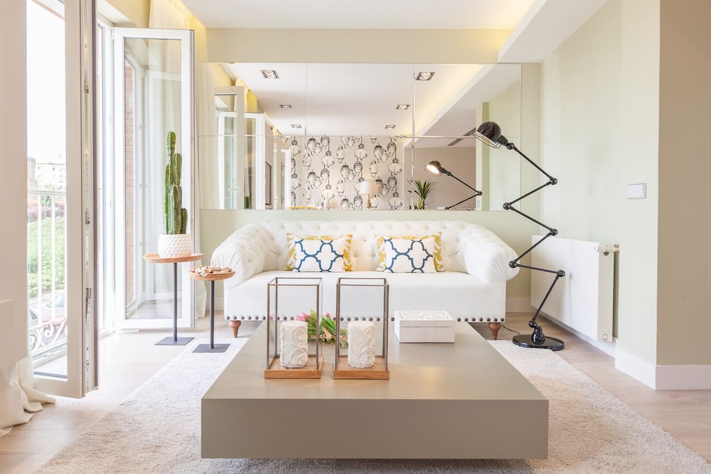 The living area has two large French windows and is furnished with a 3-seater-sofa, an armchair, a coffee table, 2 decorative side-tables, a shelving unit and a flat screen TV.