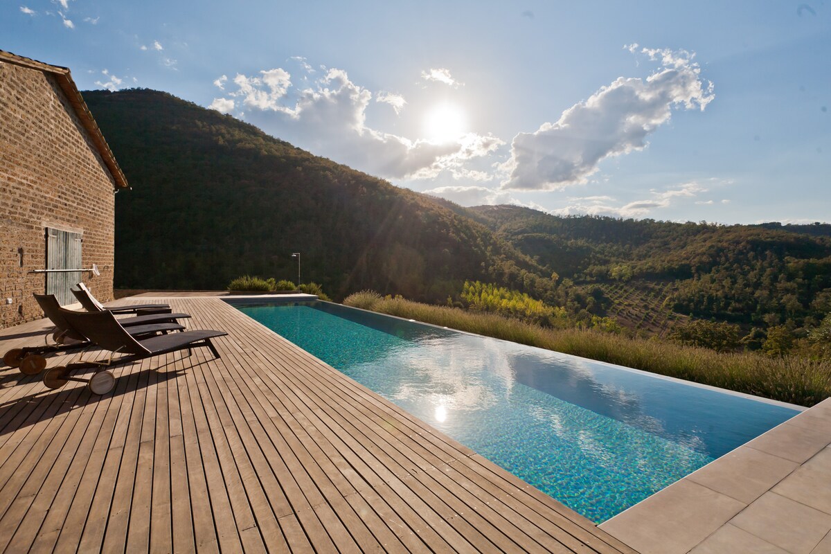 An infinity pool extends alongside a wooden deck, reflecting the sunlight as it sets behind the hills. Lounge chairs are positioned at the pool's edge, with a backdrop of lush green landscape and majestic mountains, creating a serene and tranquil environment.
