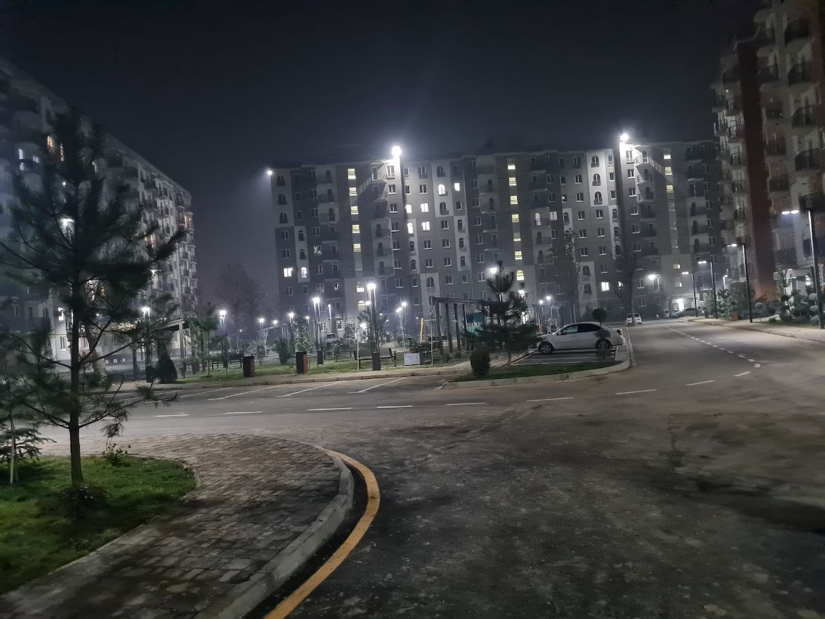 An evening view of a residential area features well-lit pathways and buildings, creating a safe and welcoming ambiance. Surrounding greenery includes small trees and a playground, contributing to the neighborhood's charm. Vehicles are parked along the curved road, indicating an active community space.