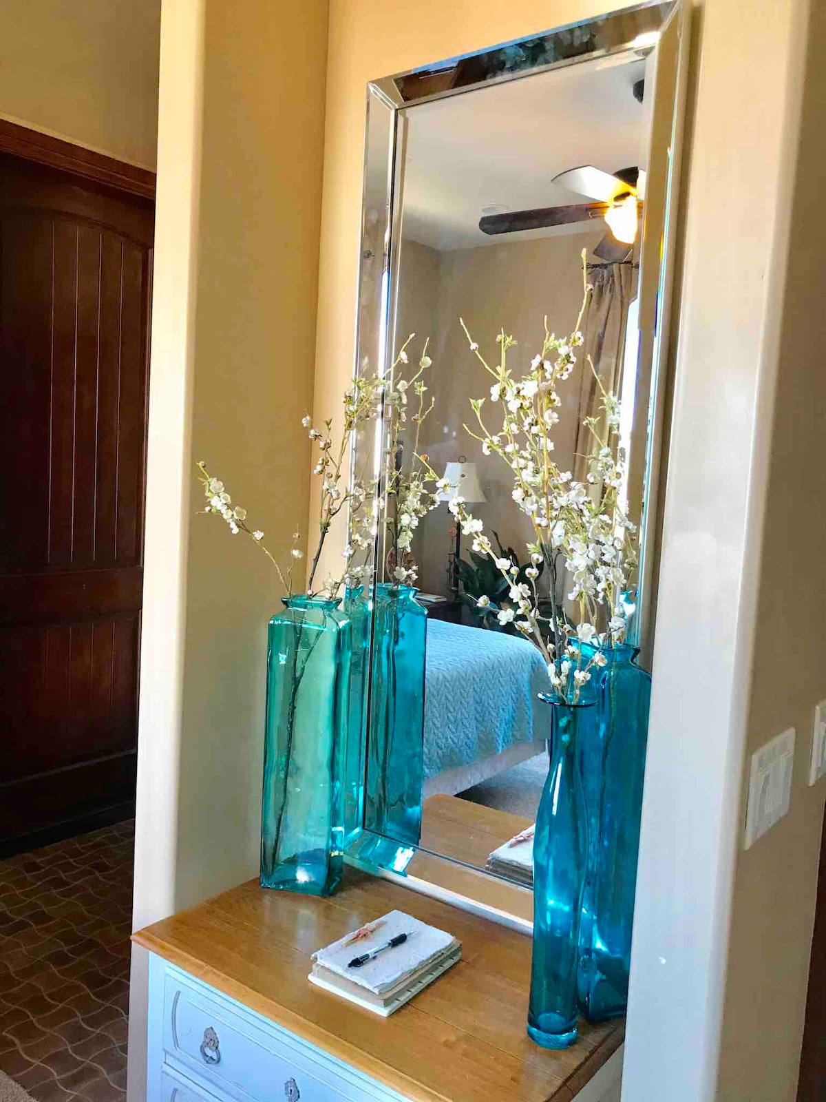 A tall, rectangular mirror reflects the softly lit room, while two vibrant blue vases hold delicate white floral arrangements. A small wooden table below features a notepad and pen, complemented by a textured background that leads toward the bedroom area.