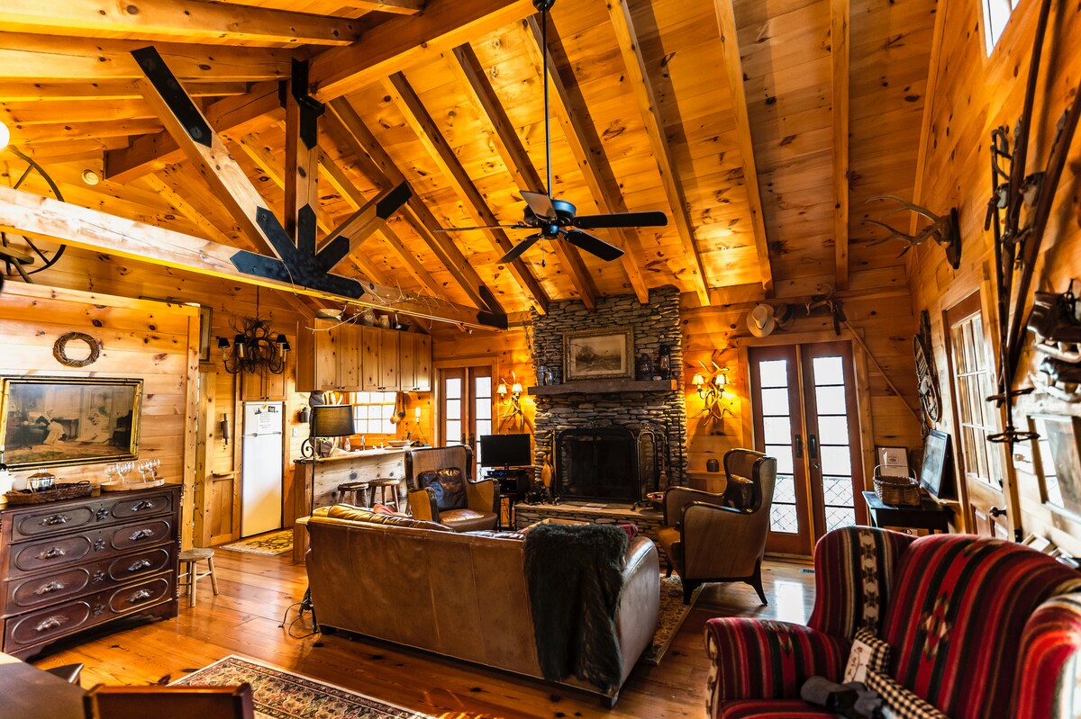 A spacious living area features a high, wooden ceiling with exposed beams. Natural light streams in through large windows, highlighting a stone fireplace as the focal point. Comfortable seating includes a leather sofa and a striped armchair, complemented by wooden furniture and rustic decor.