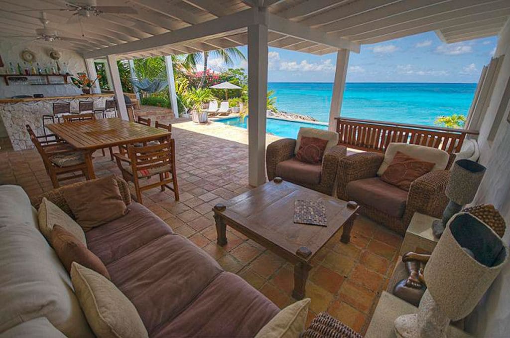 An open-air seating area features a wooden table surrounded by several dining chairs. A comfortable sofa and armchairs are arranged for relaxation, with decorative cushions. The space is bathed in natural light, offering breathtaking views of the turquoise sea through large glass openings.