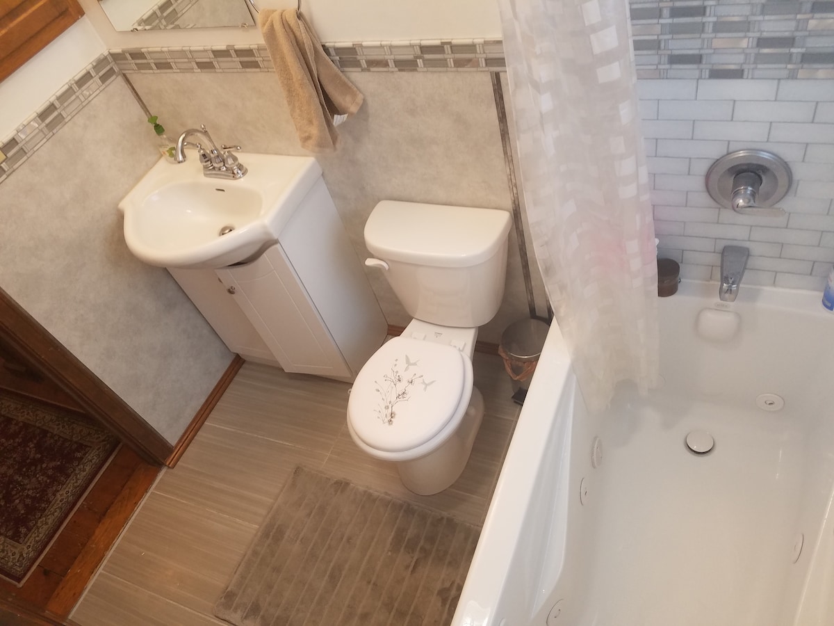 A compact bathroom features a white sink and toilet, both positioned against a pale gray tiled wall. A bathtub with a shower attachment is present, accompanied by a light curtain. A small rug lies on the floor, providing a touch of warmth to the space.